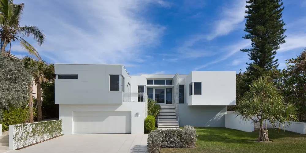 The front of the house displays a symmetrical, minimalist design with crisp white surfaces and geometric shapes. The layout emphasizes balance, complemented by carefully landscaped surroundings.
