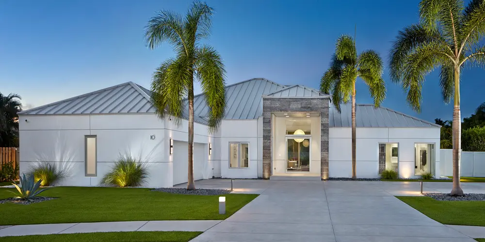 This home has undergone a remarkable transformation, moving from a dated, traditional aesthetic to a sleek and modern masterpiece. The original beige facade and shingled roof have been replaced with a clean, white finish and a contemporary metal roof. This shift not only modernizes the appearance but also introduces a sense of elegance and sophistication. Strategic lighting now highlights the home’s new streamlined architecture, enhancing both functionality and visual impact.

The mature landscaping, once giving a cozy but cluttered appearance, has been refined to feature manicured palms and a pristine lawn, framing the entrance with vibrancy and style. These changes culminate in a significant curb appeal boost, turning the residence into a standout statement of modern design and sophistication within its neighborhood. The transformation reflects a thoughtful upgrade that elevates the home to meet contemporary architectural standards while offering a welcoming and visually appealing approach.