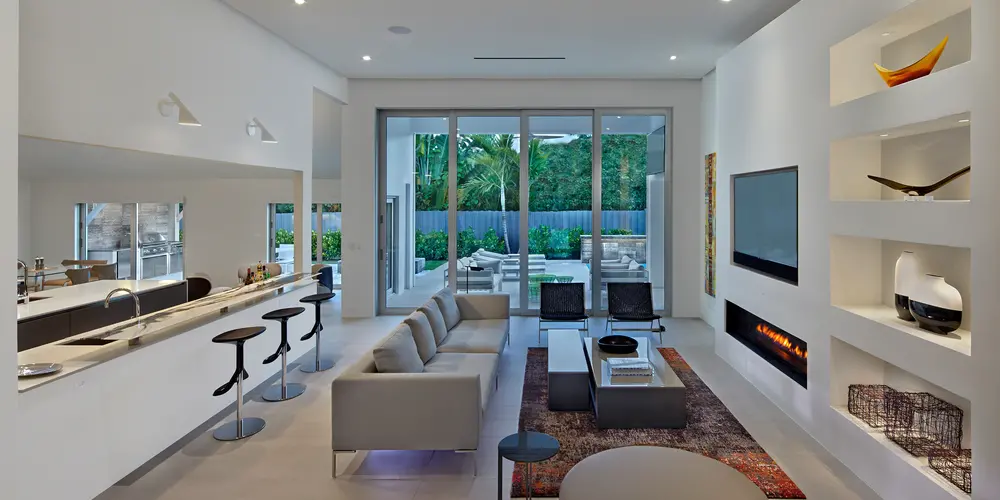 The modern living space is a harmonious blend of functionality and refined aesthetics, characterized by an open-plan layout and sleek, contemporary design. Clean lines, neutral tones, and strategically placed furnishings emphasize a minimalist approach, while built-in features like the recessed fireplace and display niches add sophistication and practicality. Large glass doors extend the living area outdoors, enhancing the sense of spaciousness and connectivity with the surrounding environment.
