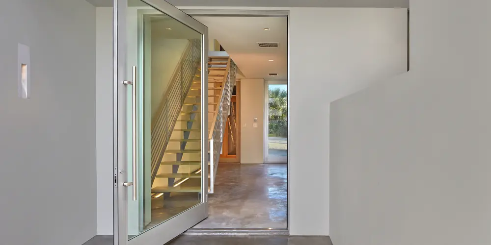 The entryway highlights contemporary design with its oversized pivot door and polished concrete flooring. The clean, uncomplicated lines invite exploration into the modern elegance of the home’s interior. The use of glass further draws natural light inside, enhancing the welcoming ambiance.

