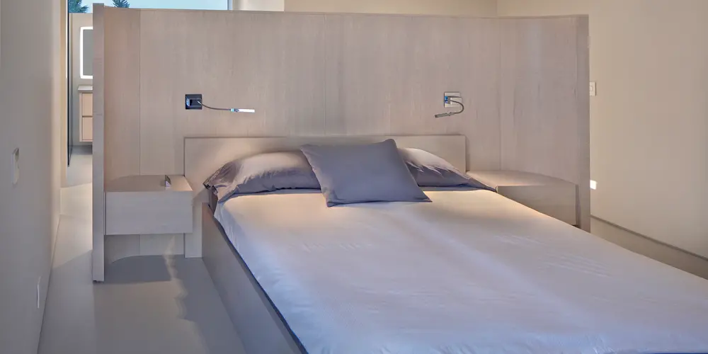 This minimalist bedroom emphasizes tranquility and functionality with a straightforward approach. The streamlined bed with integrated lighting and built-in side tables aligns with the neutral color tones to create a serene sleeping environment.

