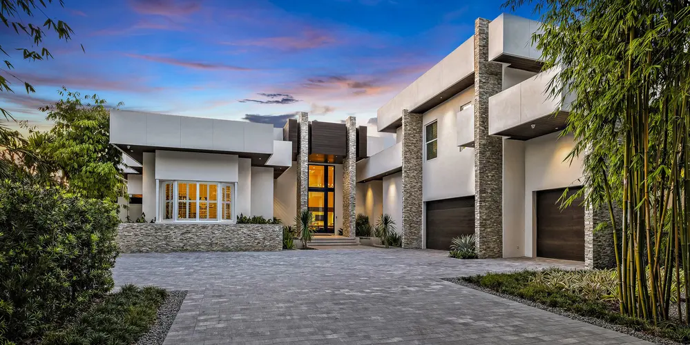 The approach to the home features a spacious driveway with intricate stonework and lush landscaping. The home's entrance is framed with large stone pillars, complementing the contemporary architecture with natural texture.