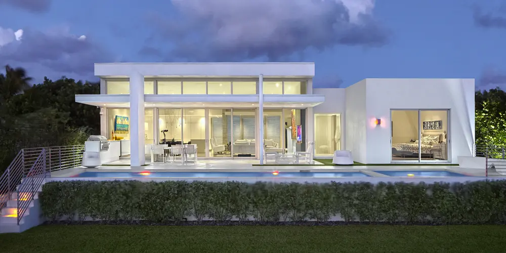  This rear view of the home at dusk showcases a stunning combination of clean lines and expansive glass surfaces. The illuminated interiors highlighted through large windows create a vibrant connection between the inside and the spacious terrace outside. The design emphasizes seamless indoor-outdoor living, enhanced by a long, sleek pool and a jacuzzi that embody luxury and relaxation.