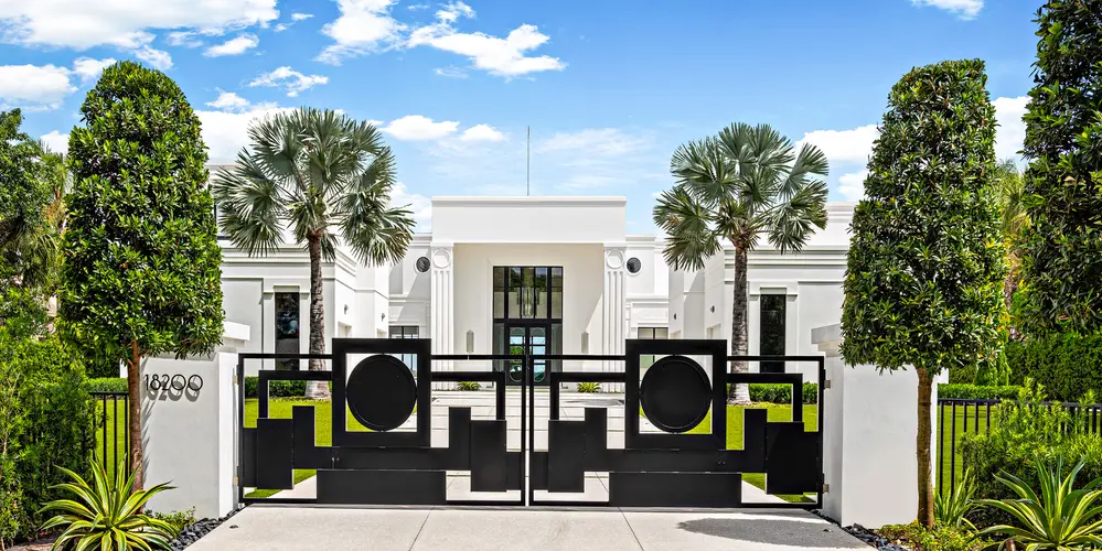 The entrance of this property exudes a sense of grandeur and exclusivity, marked by a striking geometric designed gate that perfectly complements the modern architectural style of the home. The gate, made of sleek black metal, stands in stark contrast to the white exterior of the house, creating a visually captivating focal point upon arrival. The lush greenery, featuring palm trees and meticulously pruned hedges, frames the approach and enhances the inviting ambiance of the entrance.