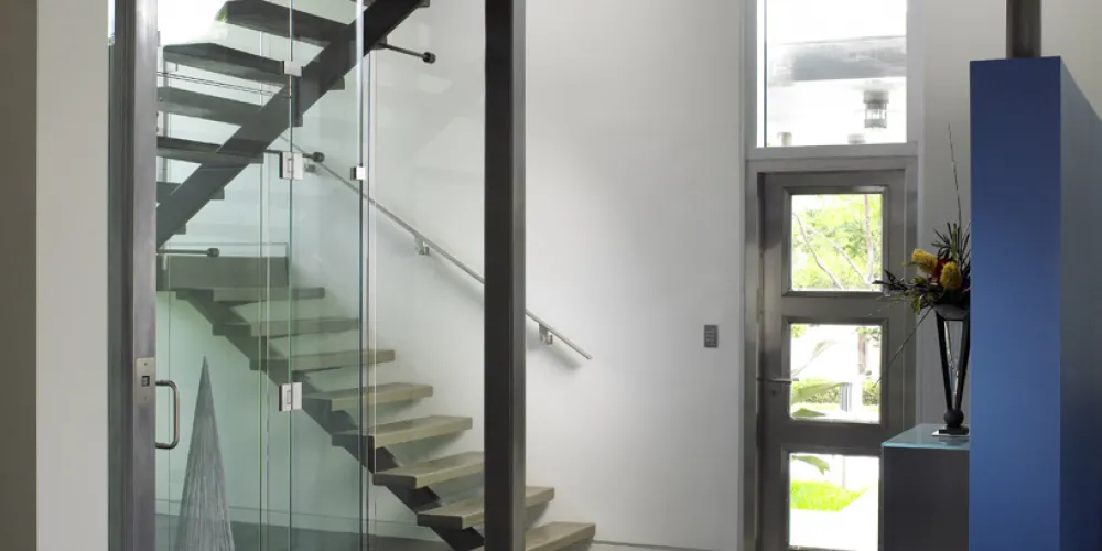 This interior view showcases a modern staircase with an elegant glass enclosure that adds depth and dimension to the space. The exposed steel beams highlight a blend of industrial design and modern minimalism. Natural light streams through a large, vertical window, emphasizing the concrete floor's clean lines and subtle textures.