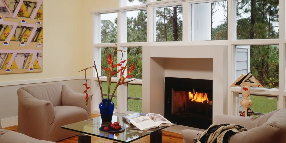 This cozy living room, framed by large windows, draws the outdoors inside, offering a view of surrounding natural beauty. Comfortable seating encircles a sleek fireplace, creating an inviting atmosphere perfect for intimate gatherings or relaxation. The soft decor and artistic touches complete the peaceful ambiance.

