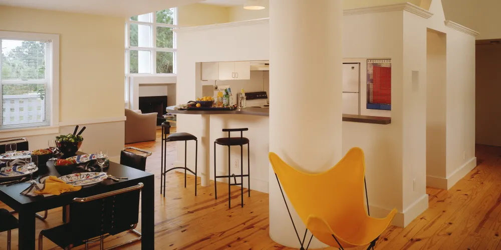 The open-concept layout fluidly connects the kitchen, dining, and lounge areas. A prominent central column divides spaces with architectural flair, while natural wooden floors and white walls maintain simplicity. A pop of color from a cheerful yellow chair enlivens the space, while the abundance of natural light enhances its airy feel.

