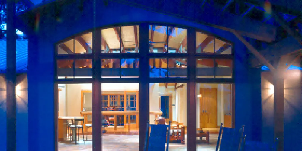 This patio creates an inviting atmosphere with its open-beamed ceiling and ambient lighting, styled to create a perfect blend of modern and rustic design elements. The glass windows allow for an expansive visual connection to the indoor area, enhancing the house's integrated living spaces.