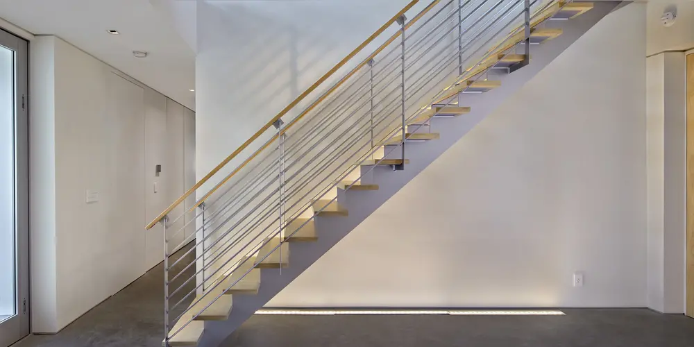 The staircase’s sleek design features open risers and a simple handrail that reflects contemporary minimalism. The use of wood and metallic finishes provides visual contrast and texture, offering an elegant yet functional pathway through the house.
