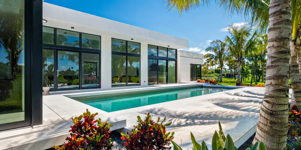 This design takes a minimalist approach with sharp lines and expansive glass, reflecting a harmonious blend of indoor and outdoor spaces. The vibrant green of the grass juxtaposes against the sleek white tiles, adding texture and contrast to the overall scene. The lush palm trees and colorful foliage complement the architectural simplicity, offering a luxurious space for relaxation and leisure.