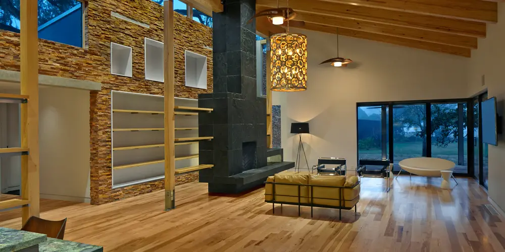 This image captures the essence of a modern, open-concept living area, elegantly balancing form and function. The space is dominated by natural wood elements, with the polished wooden floors and exposed ceiling beams evoking a sense of warmth and inviting ambiance. This earthy aesthetic is complemented by a variety of textures, from the rough stone wall feature to the smooth black stone fireplace, each contributing to the room's character and depth.