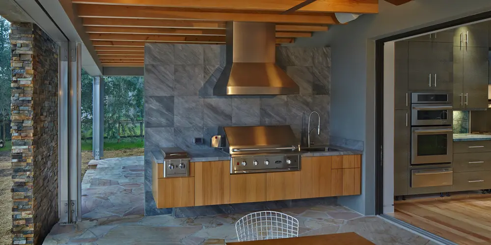This image highlights a modern outdoor kitchen area that gracefully extends the interior living space into the open air, epitomizing the seamless blend of nature and comfort. The space is designed to be both functional and aesthetically pleasing, with sleek and modern stainless steel appliances complementing natural wood cabinetry, creating an inviting cooking environment.
