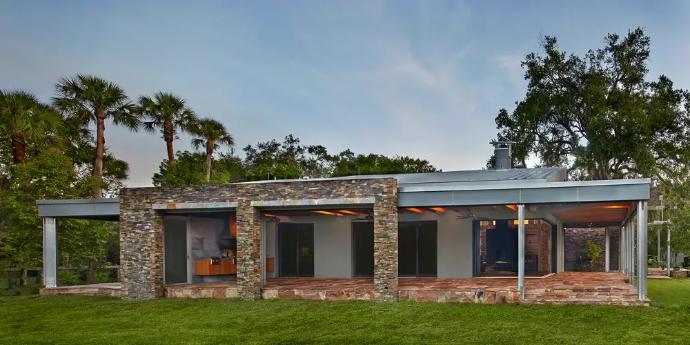 This image presents an exterior view of a contemporary residence characterized by its harmonious integration with the lush surrounding landscape. The building's facade prominently features a robust, textured stone finish that grounds the structure in its natural setting, providing a rustic charm that contrasts with the sleekness of modern design elements.