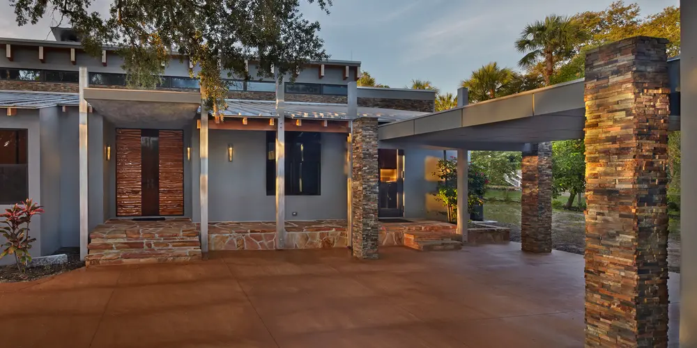 The entrance is framed by sturdy stone pillars supporting a prominent overhang, providing both protection and an eye-catching architectural detail. The stone used for the columns contrasts beautifully with the smooth, terracotta-hued concrete driveway, tying the natural elements into the home's design.

The front facade boasts clean lines and a blend of minimalist style with natural textures, creating a visually appealing contrast between the industrial elements and the warmth of natural materials. The use of large windows allows ample natural light to flood the interior, enhancing visibility and creating a seamless indoor-outdoor connection.

The overall design of this entrance combines practicality with aesthetic sophistication, setting the tone for the contemporary interior that awaits inside. The integration of natural elements with modern design principles makes this entrance both functional and aesthetically pleasing.