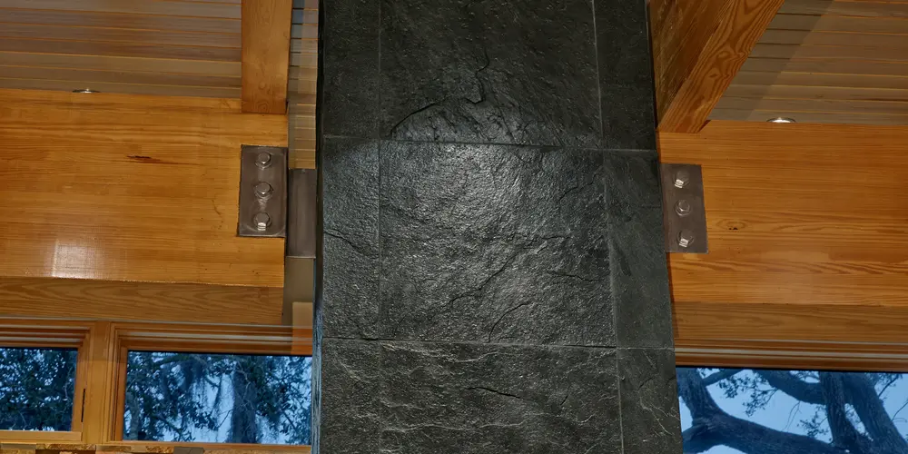 The focal point is a dark stone column that exhibits a rugged, natural appearance, emphasizing earthy textures in contrast to the surrounding smooth wooden ceiling. The stone's rich, deep color adds a dramatic tone to the interior design, grounding the space with its solidity and strength.