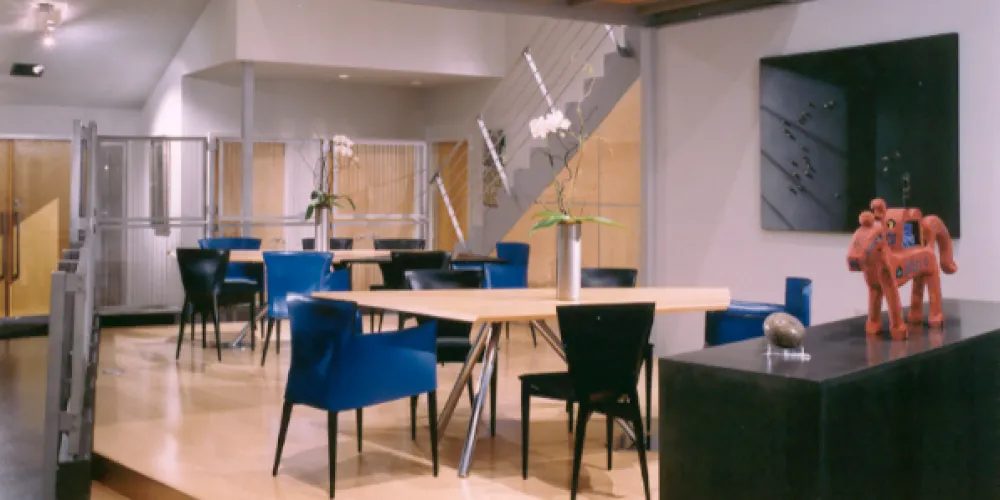 This modern dining area features a raised platform that creates a distinct zone within the open-plan space. Sleek blue chairs and a minimalist dining table add vibrancy against the clean lines of the design. The mezzanine level above makes efficient use of vertical space while the railing design provides an industrial touch.