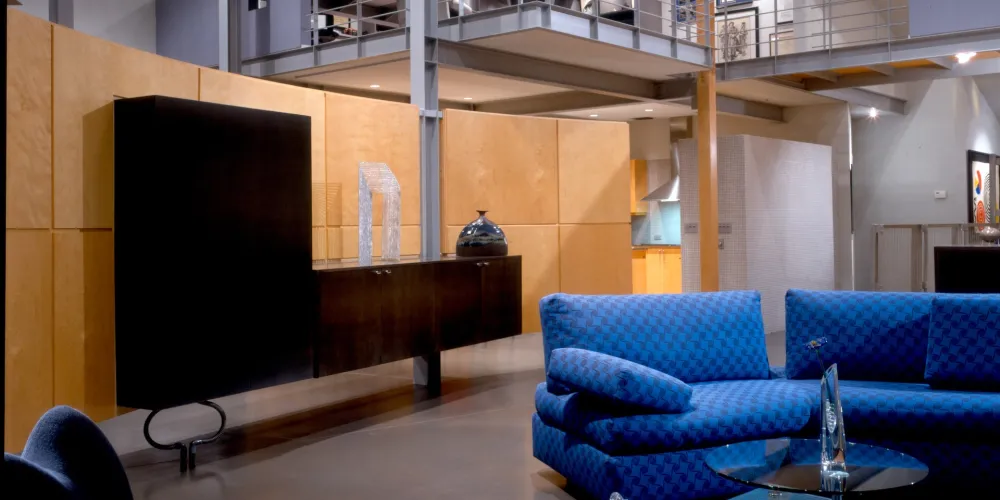 The open living area blends modern furniture design with thoughtful use of textures and color. Plush, blue seating and a glass coffee table create a cozy, inviting environment. The overhead mezzanine and open railings expand the spatial dynamic, accentuated by ambient and natural lighting.
SEO Description: Inviting living space with plush blue seating and glass coffee table, enhanced by a mezzanine that broadens the spatial appeal with modern elegance.