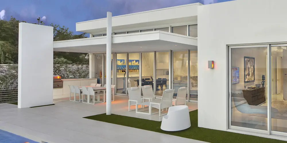 The outdoor dining and lounge area seamlessly connects with the interior, showcasing stylish furniture and ample space for entertaining. Flanked by tall glass doors, this area provides an inviting spot for alfresco dining, enhanced by the surrounding modern design and lush green touches.
